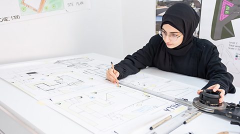 A woman wearing a black hijab working on a sketch on a desk with lots of paper and architectural blueprints.