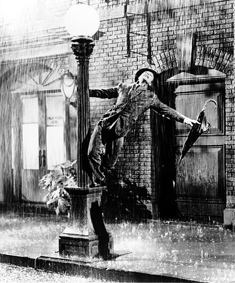 Black and white photo of actor Gene Kelly singing and dancing around a lamppost in simulated rain, an umbrella in one hand