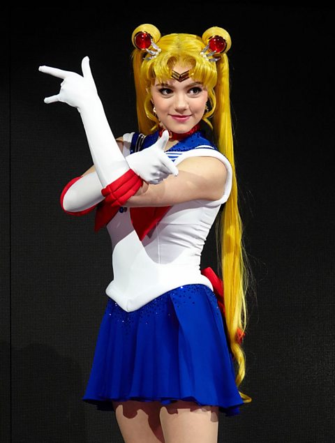 Russian figure skater Evgenia Medvedeva wears a Sailor Moon costume of a long blonde wig, in pigtails, a white top, blue skirt, white gloves and red detailing.