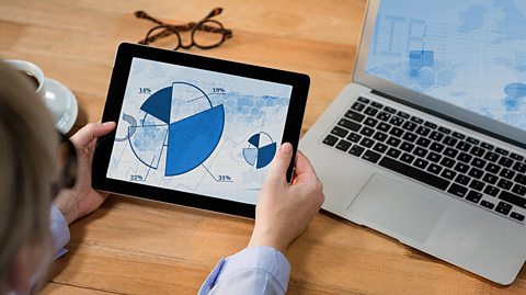 A white person holding a tablet with a pie chart on it. Behind is an open laptop. 