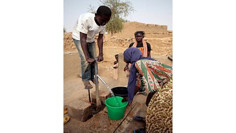 A young boy operating a standpipe. Water pours from the standpipe into a bucket held by a woman.