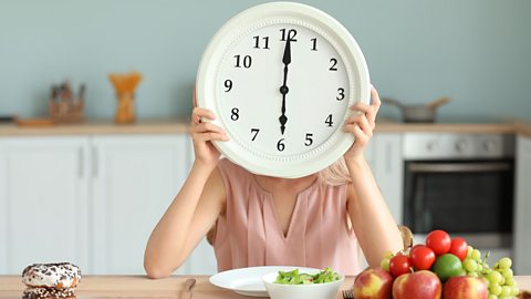 A child sits at the dinner table with food in front of her and holding a big clock in front of her head which shows the time is six o'clock