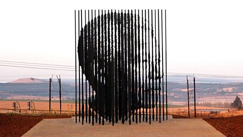 A group of fifty black columns with uneven edges. The side profile of Nelson Mandela can be seen in the shape they form. Behind the statue is an empty landscape of sandy fields.