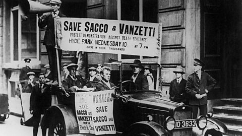 A group of men travel in a car, while one man shouts through a megaphone. The car is covered in 'Save Sacco and Vanzetti' banners