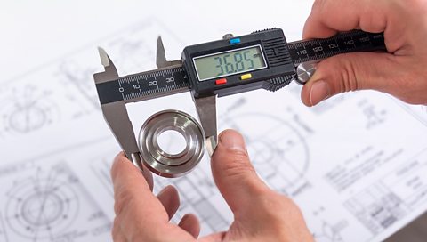 A digital vernier caliper - a measuring device with a black handle and a digital reading - being held above some paper blueprints.