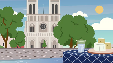 A cartoon of Notre Dame cathedral. It is tall with two towers and arched windows. It is in between two trees. At the front of the cartoon is a table with some cheese and a drink on it.