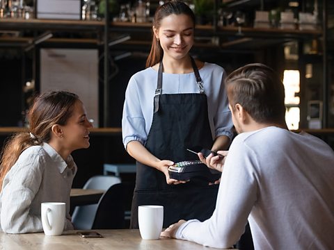 A white waitress smiling at two customers as one of them taps their card on a card machine to pay. They have two coffee cups next to them.