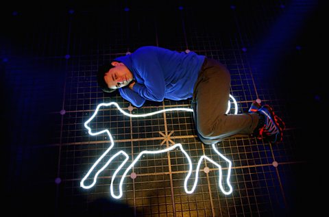 A young man lies alongside the illuminated outline of a dog on the floor. Part of a stage production