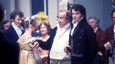 An image of people dressed in period style dresses and suits at a ball. In the centre of the picture, there is a tall, dark haired man, scowling towards the the camera, and a portly elderly man laughing