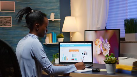 A black woman with a blue shirt is sat at a desk writing on a notepad. A laptop screen is open and there is a second computer screen behind.