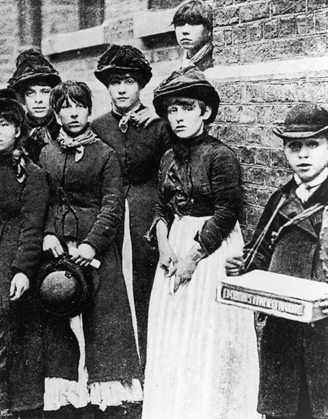 A black and white photo of a group of young women on strike. Most are wearing hats with their hair tied back, and long dresses.