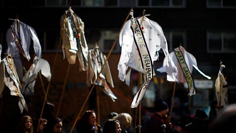 A colour photo of women holding up shirts and blouses on sticks. On each shirt is a sash with the name of a woman who died in the fire on it.