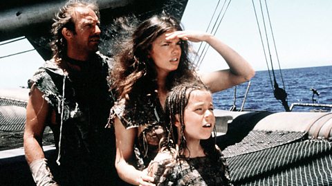 A photo from the film Waterworld. A man, woman and young girl stand on a boat looking out to sea, wearing worn clothing.