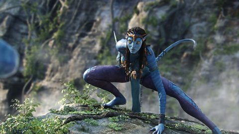 A photo from the film Avatar. A tall blue-skinned alien, with long black hair and wearing a gold circlet on her head, crouches down on an ivy-covered rock, ready to fight.