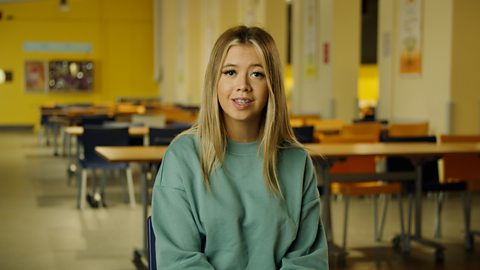 A student smiling and wearing a green sweater, sitting in front of a class room with desks.