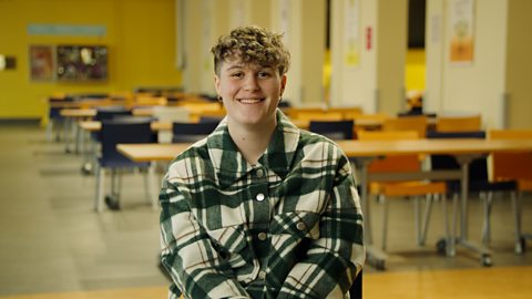 A smiling student wearing a green and cream chequered shirt, sitting in the front of a classroom.