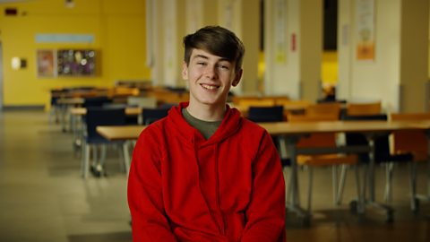 A smiling student wearing a red hoodie an sitting in front of a classroom.