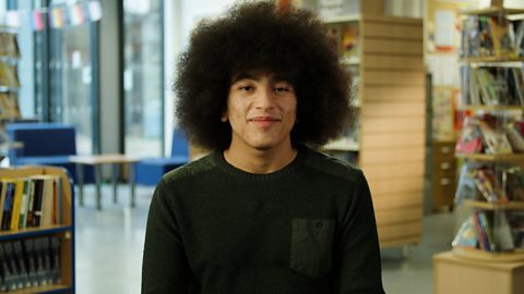 A student wearing a military green coloured jumper sits in a library, surrounded by books and bookshelves.