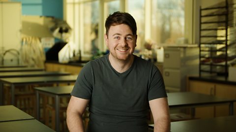 Anxiety Josh smiling, wearing a grey t-shirt and sitting in front of a classroom filled with desks and cabinets.