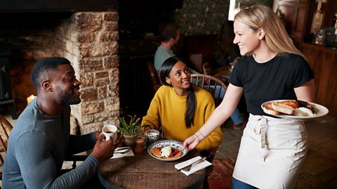 A photograph of a waiter serving food to two people at a table.