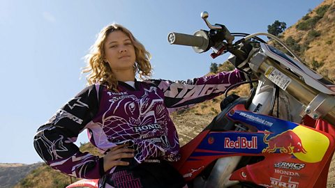 Motocross rider Ashley Fiosk sits astride a Red Bull motorbike. She is wearing purple and white leathers and has wavy blonde hair.