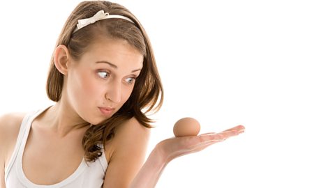A woman holds an egg in the palm of her hand looking confused by it