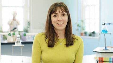 A smiling person wearing a yellow top, sitting in a science lab.