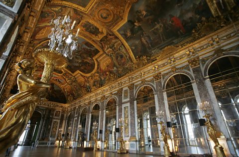 A photo of the interior of the Hall of Mirrors at the Palace of Versailles. Along the right hand wall are floor to ceiling mirrors, with tall gold candleholders between them. One of these is in the foreground, close to the camera. The ceiling features several paintings as well as extensive gold.