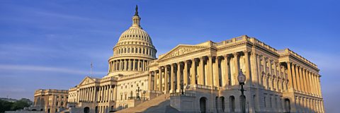 A photo of the exterior of the Capitol Building, taken from a side angle. The sky behind the building is bright blue with little cloud. The building is made of white stone with numerous columns. In the centre of the roof there is a large dome, also covered in columns.