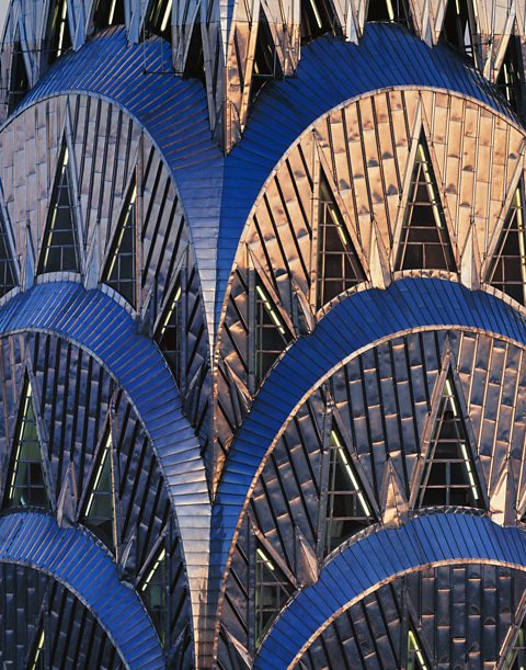 A close-up exterior photo of the top floors of the Chrysler Building. The outside of the building is covered in materials, with triangle shaped windows, positioned on top of each other in the shape of an arch.