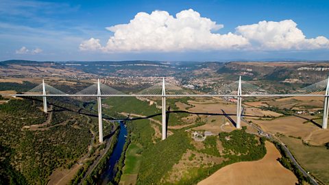 Photo of a long white road bridge supported by seven columns. Below the bridge is a river and fields.