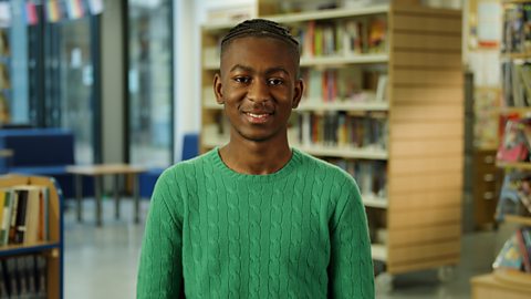 A student smiling and wearing a green sweater, sitting in front of a school library.