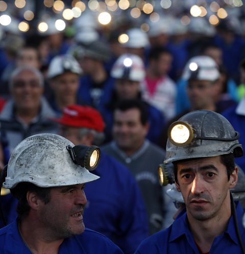 A crowd of miners wearing overalls with hard hats and the lights on their hard hats turned on.