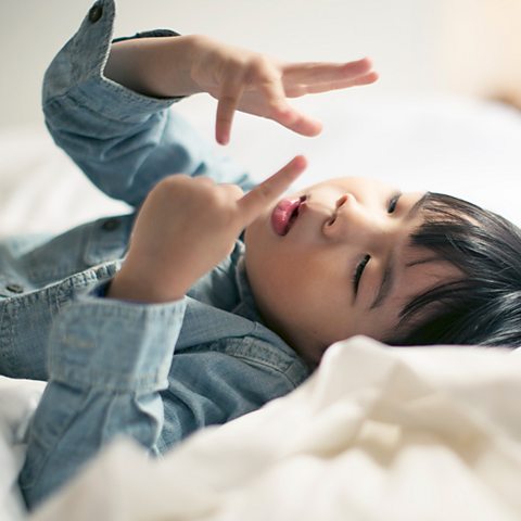 A young boy in a blue denim shirt lies on his bed and counts on his fingers. He uses both hands.