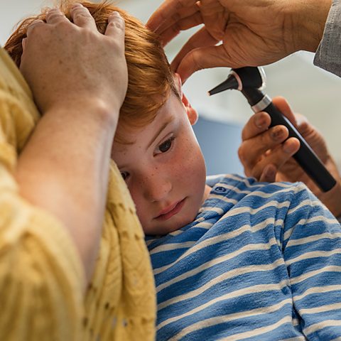 Young child is getting his ears checked by a doctor. He is being checked for Glue Ear. The boy sits on his parent's lap.