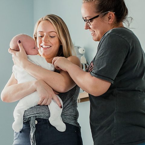 Glue Ear Two women are holding a new born baby in its bedroom. They are looking lovingly at it. It is wearing a white onesie.