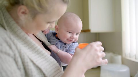 A mum is doing the washing up with her 3-month-old son in the kitchen. She is holding a plastic glass filled with water.