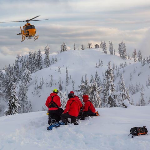 Photograph of a mountain rescue team on a snowy mountain with a rescue helicopter hovering above.