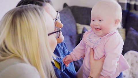 A baby and her two mums sit on their sofa. The baby is smiling as they respond to her babble.