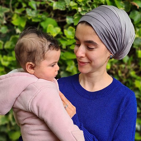 Safeeya Habaik and her baby daughter standing in front of a green hedge. Safeeya is wearing a blue jumper.