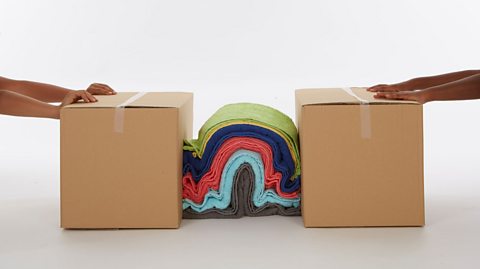 Two sets of arms each pushing a cardboard box towards each other. There is a pile of towels in between the cardboard boxes which is being pushed upwards, creating an arch.