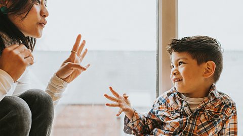 A cute young boy gestures using his hands. A woman, who sits close-by, listens and watches him intently as he explains something.