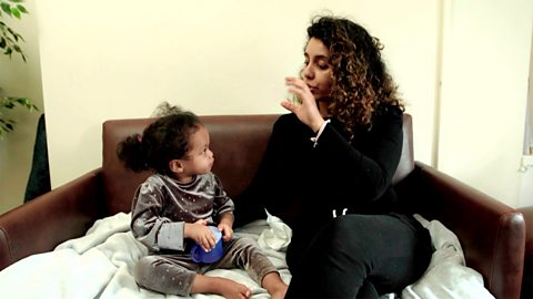 A mum and her daughter sit on the sofa. They are learning the word for "cup" while mum signs it.