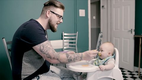 A dad feeds his son at a high chair in their kitchen. The baby is around 6 months old. Dad has tattoos down one arm and is wearing glasses.