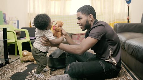 A dad and his son sit on the floor in their living room and play What's in the Bag together. Dad has pulled out a teddy.