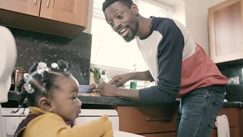 A dad is cutting food in the kitchen. His daughter sits on a high chair. He is explaining what he is doing to her.