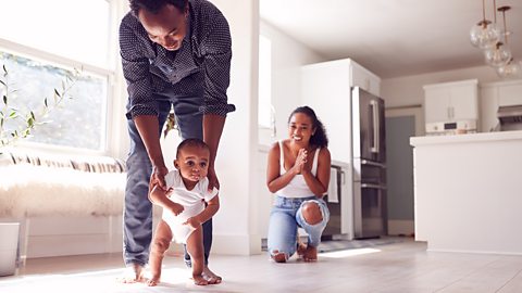 Parents Encouraging Smiling Baby Daughter To Take First Steps And Walk At ˿