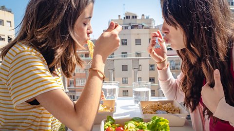 A picture of two young women eating pasta on a balcony.