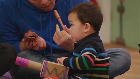 A dad and his son sit in the Toddler Club and play with a homemade cardboard box guitar and make music together.