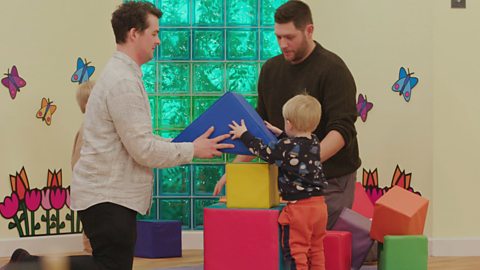 Two dad build a massive tower together with their sons. They are using soft multicoloured blocks. They are in the Toddler Club.